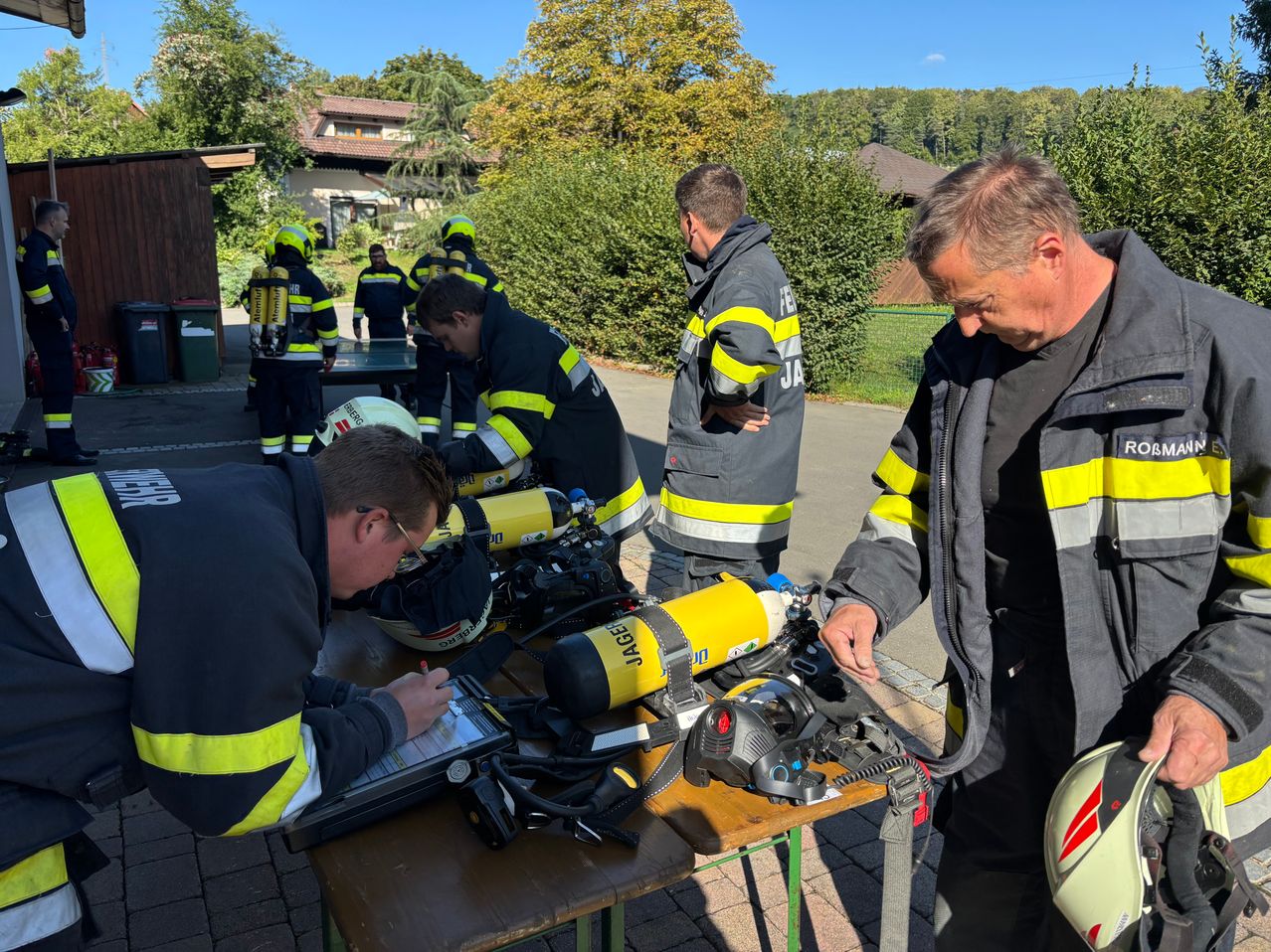 Eine Gruppe von Feuerwehrleuten ist um einen Tisch versammelt und überprüft Ausrüstung. Ein Mann schreibt auf einem Tablet, während andere Sauerstoffflaschen inspizieren.