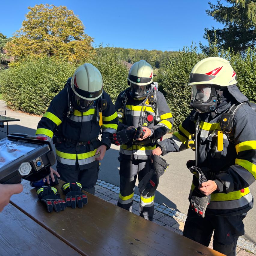 Drei Feuerwehrleute in voller Ausrüstung stehen im Freien, einer hält ein Gerät und ein anderer untersucht Handschuhe auf einem Tisch.