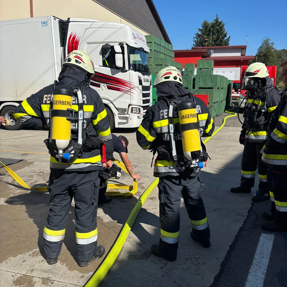 Feuerwehrleute in voller Ausrüstung, einschließlich Helmen und Sauerstoffflaschen, stehen und beobachten einen Mann, der an einem gelben Schlauch auf dem Boden arbeitet. Ein weißer LKW und grüne Kisten sind im Hintergrund.