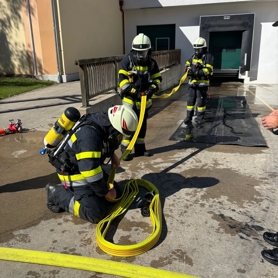 Drei Feuerwehrleute in voller Ausrüstung üben mit einer Schlauchleitung auf einer nassen Betonoberfläche. Einer kniet, ein anderer steht und der dritte hält den Schlauch.