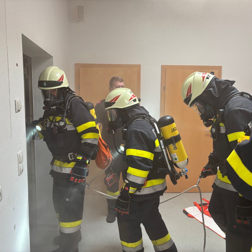 Vier Feuerwehrleute in voller Ausrüstung, einschließlich Helme und Sauerstoffflaschen, betreten einen Raum durch eine offene Tür. Einer hält eine Taschenlampe.