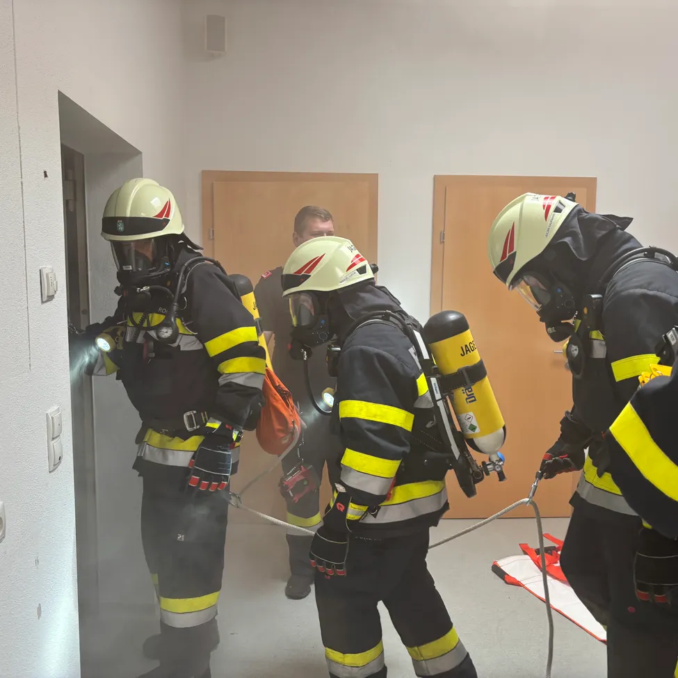 Vier Feuerwehrleute in voller Ausrüstung, einschließlich Helme und Sauerstoffflaschen, betreten einen Raum durch eine offene Tür. Einer hält eine Taschenlampe.