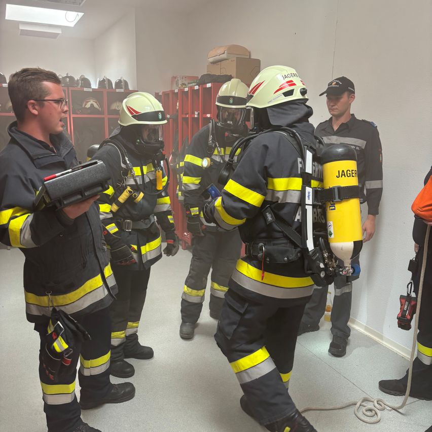 Eine Gruppe von Feuerwehrleuten in voller Ausrüstung, einschließlich Helmen und Sauerstoffflaschen, versammelt sich in einem Raum, wahrscheinlich für eine Schulung. Ein Feuerwehrmann hält ein Gerät, während ein anderer einen Hut trägt.