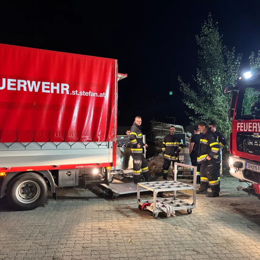 Feuerwehrleute laden nachts Ausrüstung auf einen LKW. Der LKW hat eine rote Abdeckung mit weißem Text.