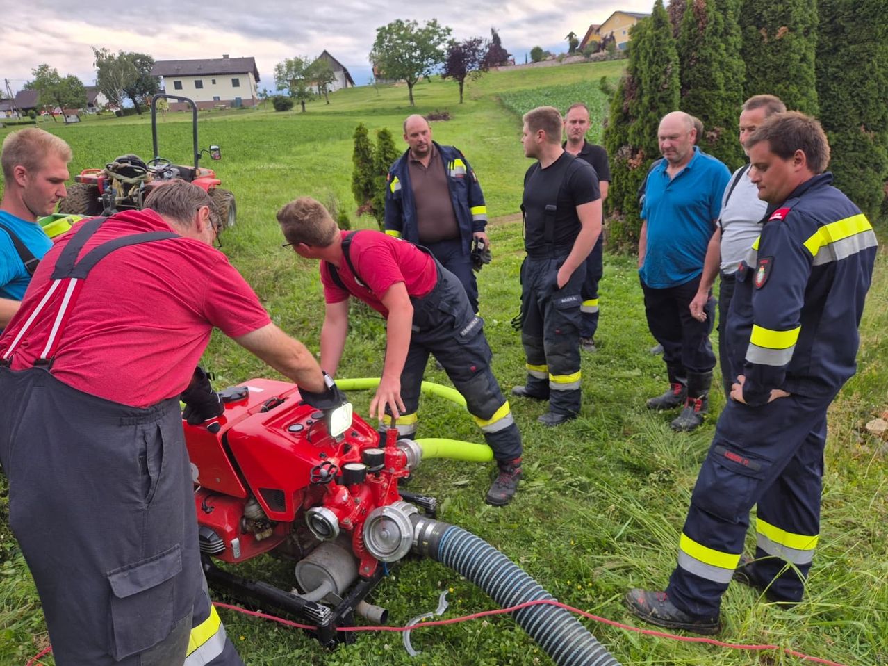 Eine Gruppe von Männern in Uniform arbeitet an einem roten Feuerwehrauto auf einem Rasenfeld. Ein Mann überprüft die Ausrüstung, während andere zusehen.
