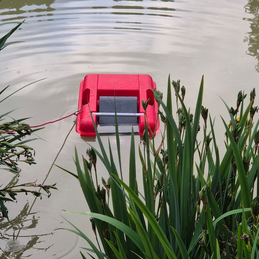 Ein rotes rechteckiges Gerät mit grauer Abdeckung schwimmt in einem Gewässer. Es ist mit einem roten Seil verbunden. Grüne Pflanzen umgeben das Gerät.