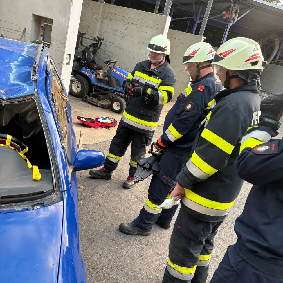 Drei Feuerwehrleute in voller Ausrüstung stehen um ein beschädigtes blaues Auto, einer hält ein Werkzeug, ein anderer untersucht das Fahrzeug. Ein roter Beutel und ein Rasenmäher sind im Hintergrund.