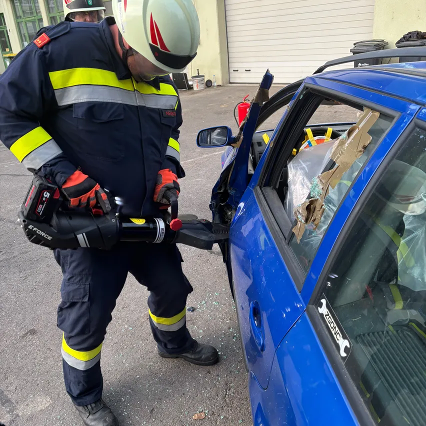 Ein Feuerwehrmann in voller Ausrüstung benutzt ein Werkzeug, um eine Autoscheibe zu zerbrechen. Das Auto ist blau und hat eine beschädigte Scheibe.