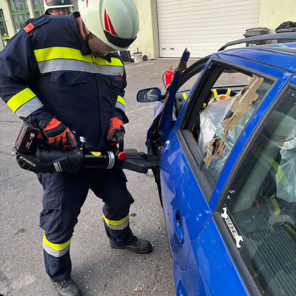 Ein Feuerwehrmann in voller Ausrüstung benutzt ein Werkzeug, um eine Autoscheibe zu zerbrechen. Das Auto ist blau und hat eine beschädigte Scheibe.
