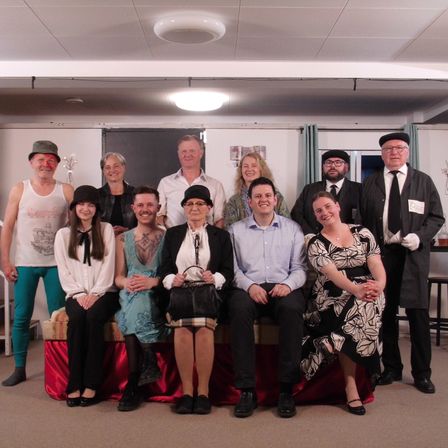 Eine Gruppe von Menschen in Vintage-Kleidung posiert für ein Foto, lächelt und steht, einige sitzen auf einer Bank. Der Raum hat eine moderne Decke und weiße Wände.