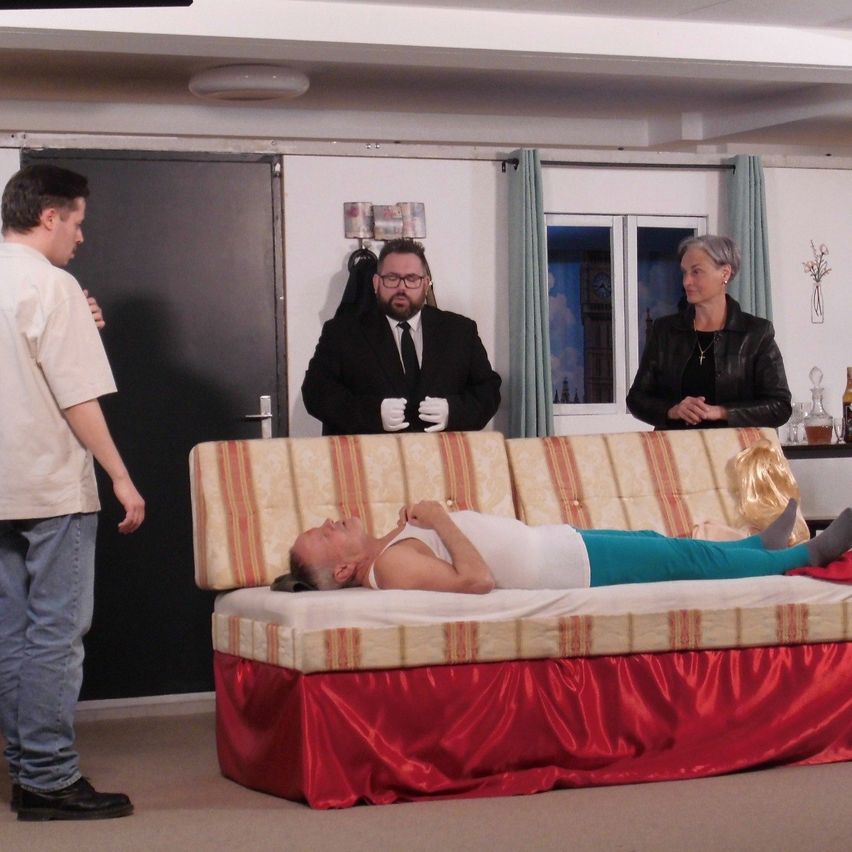 Ein Mann liegt auf einem Sofa, während ein Mann und eine Frau in formeller Kleidung hinter ihm stehen und ein anderer Mann zusieht. Im Hintergrund sind eine Uhr und Blumen zu sehen.