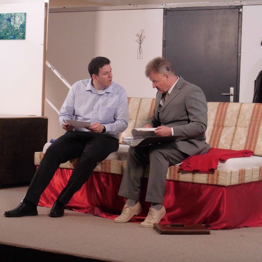 Zwei Männer sitzen auf der Bühne, einer liest ein Papier, der andere hält ein Papier, beide tragen Anzüge. Hinter ihnen ist ein Sofa mit rotem Stoff und eine Tür.