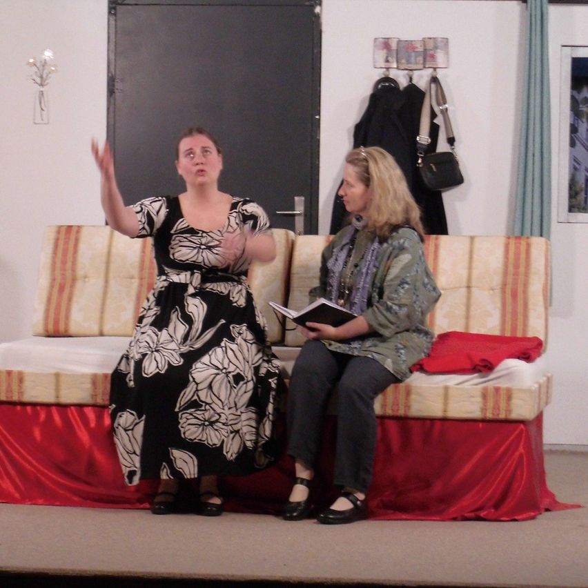 Zwei Frauen sitzen auf einer Couch auf der Bühne, eine trägt ein schwarzes Blumenkleid und die andere eine grüne Jacke. Dahinter ist eine Tür leicht geöffnet, und eine schwarze Tasche hängt an der Wand.