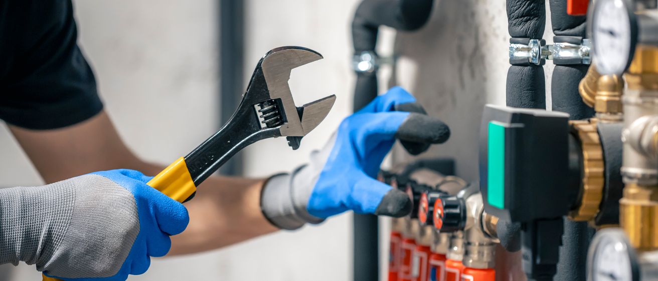 Ein Arbeiter mit Handschuhen benutzt einen Schraubenschlüssel an einem Ventil, nahe an Gasflaschen an einer Wand.