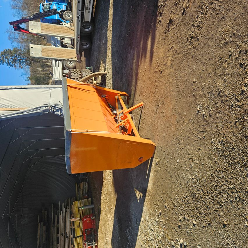 Ein orangefarbener Schneepflug steht am Rande einer unbefestigten Straße. Dahinter ist ein Lkw mit Holzbrettern beladen.