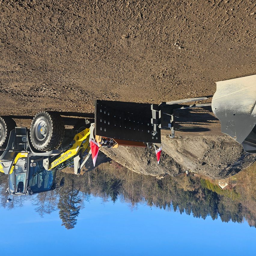 Ein gelber Baufahrzeug wird benutzt, um in einen Hang zu graben, mit einer Reflexion des blauen Himmels und der Bäume.