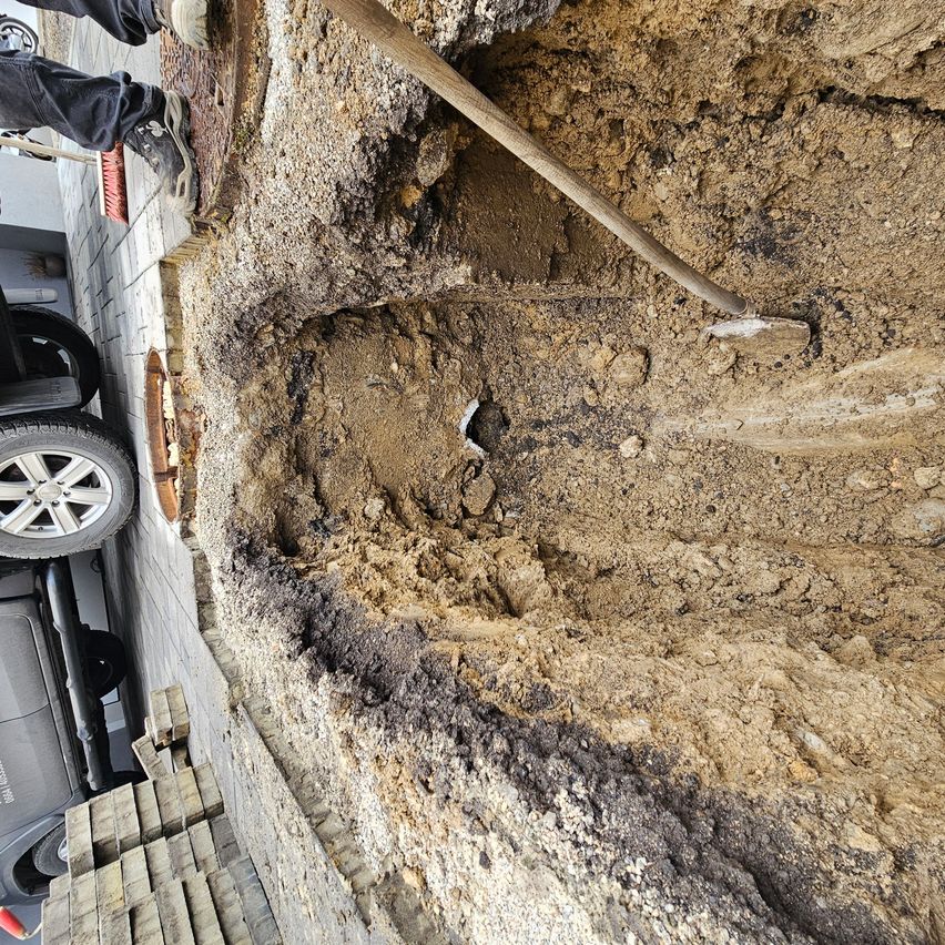 Eine Person steht neben einem Auto und gräbt mit einem Holzspaten in die Erdoberfläche. Lockerer Boden und Steine umgeben das Loch.