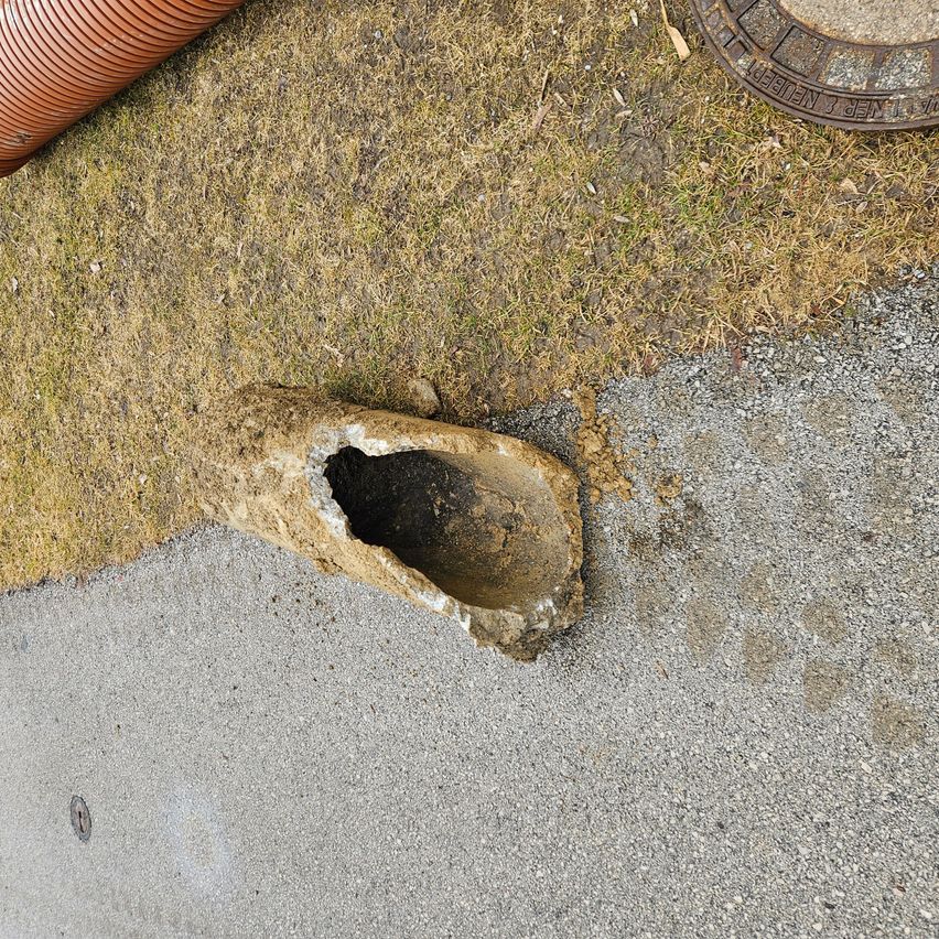 Ein Luftbild eines zerbrochenen Rohres am Straßenrand, mit trockenem Gras, einem Kanaldeckel und einem Schlauch in der Nähe.