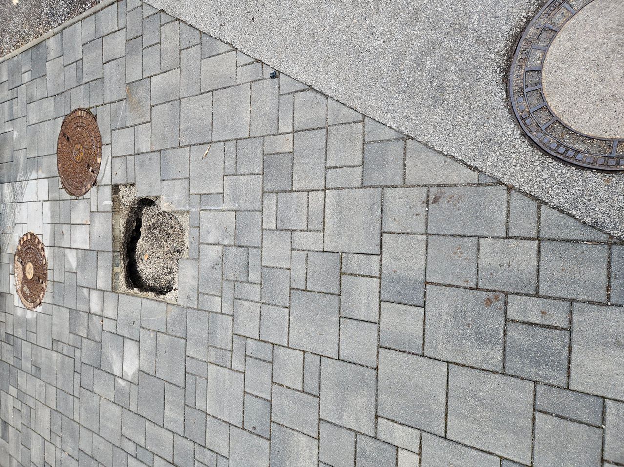Ein Blick von oben auf einen Bürgersteig mit einer beschädigten Stelle. Der beschädigte Bereich ist mit Kies und Schutt gefüllt. Zwei Schachtdeckel sind auf dem Bürgersteig sichtbar.