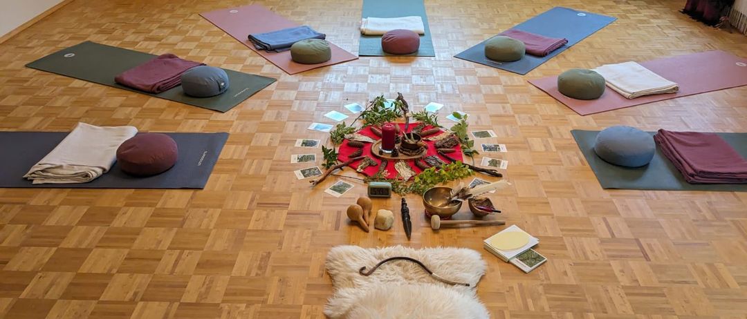 Ein Raum mit Holzboden und in einem Kreis angeordneten Yoga-Matten, einem weißen Pelzteppich, einer Kerze und einer Sammlung von Karten auf einem Tisch. Stühle stehen in der Nähe des Fensters.