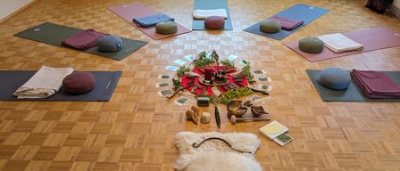 Ein Raum mit Holzboden und in einem Kreis angeordneten Yoga-Matten, einem weißen Pelzteppich, einer Kerze und einer Sammlung von Karten auf einem Tisch. Stühle stehen in der Nähe des Fensters.