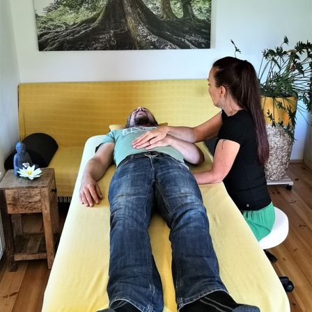 Ein Mann liegt auf einem gelben Bett und bekommt eine Massage von einer Frau in einem schwarzen Shirt. Hinter ihnen steht eine Couch mit einem Kissen und einer Pflanze. Es gibt einen Tisch mit einer weißen Blume und ein Bild von Wurzeln an der Wand.