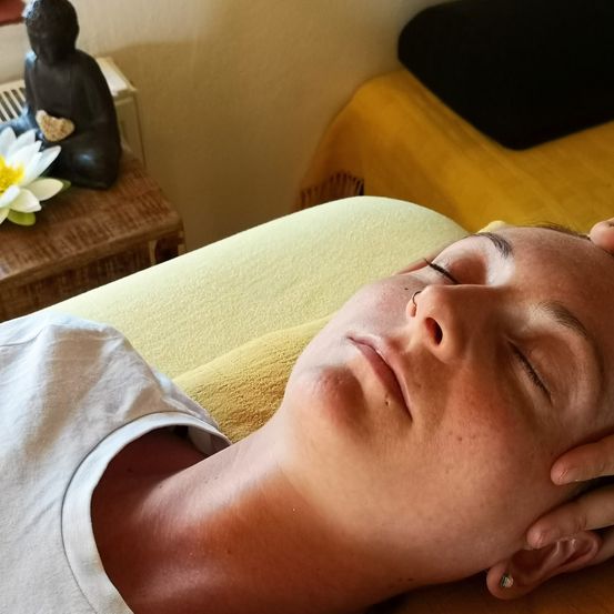 Eine Person liegt auf einem gelben Kissen, die Augen geschlossen, und erhält eine Massage. Eine weiße Blume und eine schwarze Buddha-Statue befinden sich auf einem nahegelegenen Tisch.