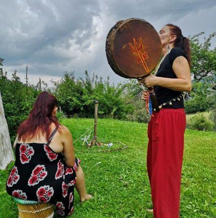Zwei Frauen befinden sich auf einer Wiese. Eine steht und hält eine Trommel mit einem Dreizack-Symbol, während die andere sitzt und zuschaut. Im Hintergrund sind Bäume und Büsche zu sehen.