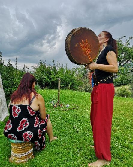 Zwei Frauen befinden sich auf einer Wiese. Eine steht und hält eine Trommel mit einem Dreizack-Symbol, während die andere sitzt und zuschaut. Im Hintergrund sind Bäume und Büsche zu sehen.