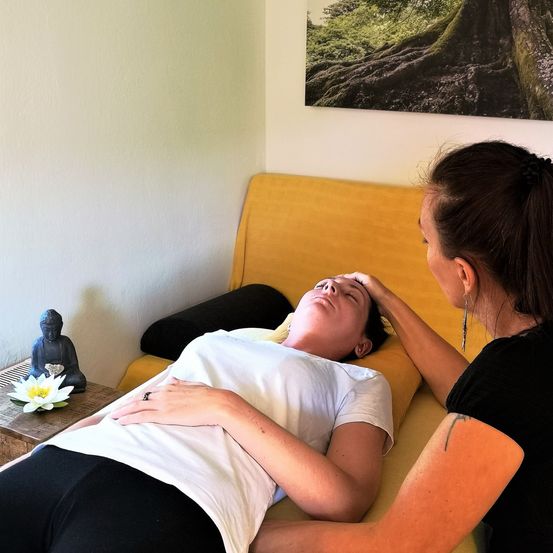 Eine Frau erhält eine Gesichtsmassage von einer anderen Frau in einem Raum mit einem Baumfoto an der Wand.