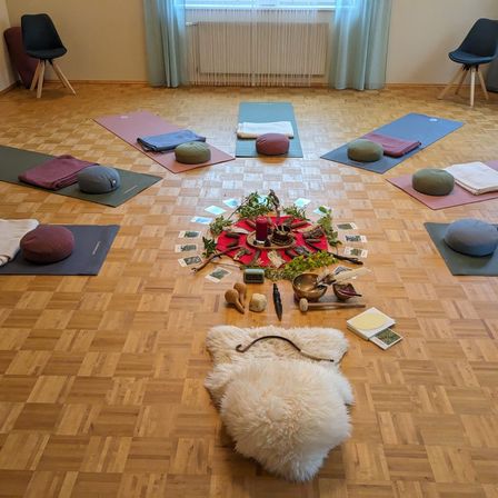 Ein Yogastudio mit Holzboden, mehreren Yogamatten und einem zentralen Bereich mit Kerzen, Pflanzen und verschiedenen Gegenständen, die um ein rotes Tuch herum angeordnet sind. Stühle stehen in den Ecken.