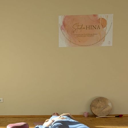 Eine Person liegt auf einer Yogamatte in einem Raum mit einer beigefarbenen Wand, einem Poster, einer Trommel und einer Tasse. Eine Steckdose befindet sich an der linken Wand.