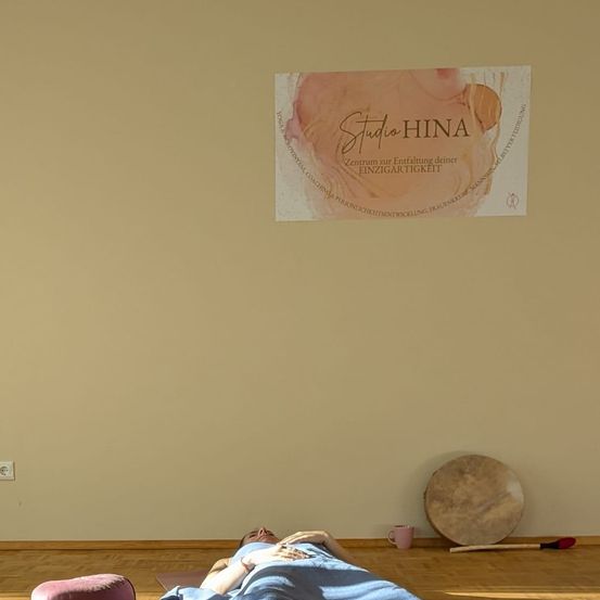 Eine Person liegt auf einer Yogamatte in einem Raum mit einer beigefarbenen Wand, einem Poster, einer Trommel und einer Tasse. Eine Steckdose befindet sich an der linken Wand.