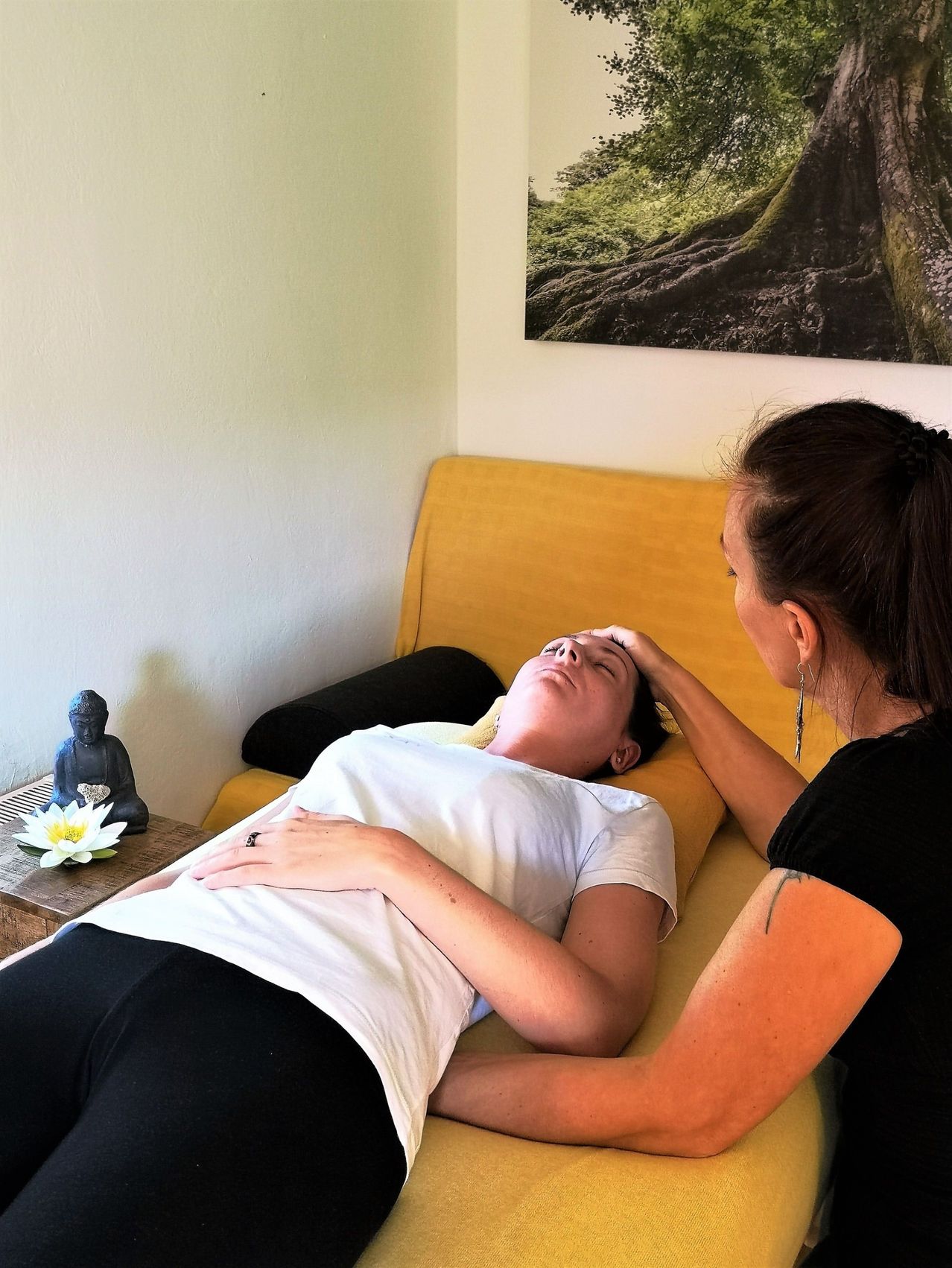 Eine Frau liegt auf einem gelben Sofa und erhält eine Massage von einer anderen Frau. Eine Buddha-Statue und eine weiße Blume stehen auf einem Tisch neben dem Sofa. Ein großes Bild eines Baumes hängt an der Wand.