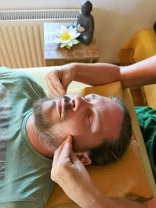 Ein Mann liegt auf einem Bett mit einem Kissen und erhält eine Gesichtsmassage. Eine andere Person massiert sein Gesicht. Ein kleiner Tisch mit einer Blume und einer Statue steht hinter ihnen.