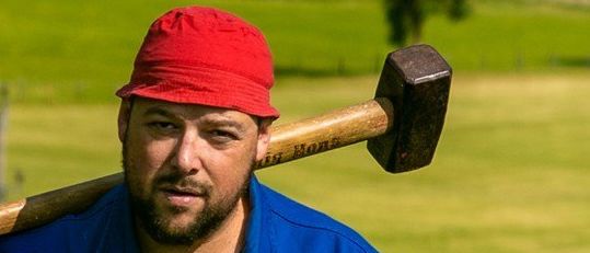 Ein Mann mit rotem Hut und blauem Shirt hält einen großen Hammer auf einem Grasfeld.