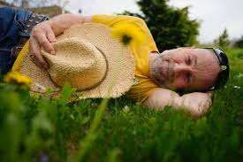Ein Mann liegt auf einem Grasfeld, trägt ein gelbes Shirt und einen Strohhut. Er hat eine Sonnenbrille auf der Stirn und scheint sich auszuruhen.