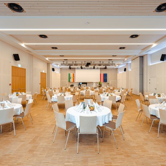 Ein Konferenzsaal mit mehreren runden Tischen und Stühlen, arrangiert für eine formelle Veranstaltung. Der Raum hat eine Bühne mit einer Projektionsleinwand und Flaggen. Der Boden ist aus Holz und die Wände sind weiß.