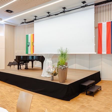 Eine leere Bühne mit Klavier, Flaggen und Topfpflanzen vor einer Holzwand und einer weißen Leinwand.