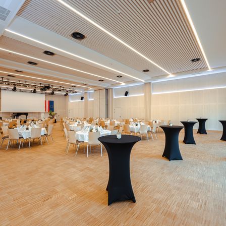 Ein Konferenzsaal mit Tischen und Stühlen für eine Veranstaltung vorbereitet. Der Raum hat Holzböden und weiße Wände. Es gibt Flaggen an der Wand.