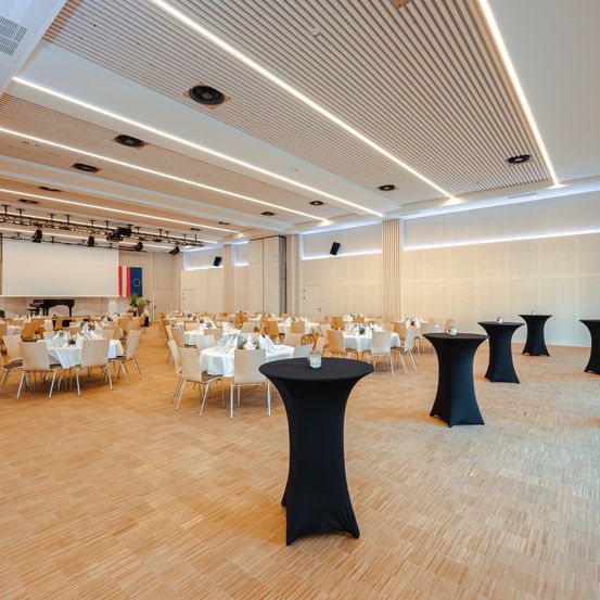 Ein Konferenzsaal mit Tischen und Stühlen für eine Veranstaltung vorbereitet. Der Raum hat Holzböden und weiße Wände. Es gibt Flaggen an der Wand.