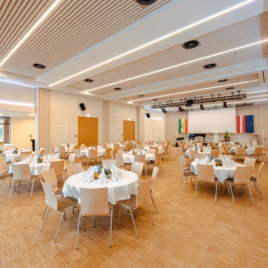Ein Bankettsaal mit Holzböden und weißen Wänden, gefüllt mit runden Tischen, die mit weißen Tischdecken und Blumen sowie Servietten bedeckt sind. Flaggen verschiedener Länder sind an der Wand angebracht.