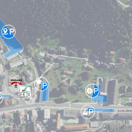 Eine Luftaufnahme zeigt eine kleine Stadt mit mehreren Häusern und Gebäuden. Parkzeichen und eine Box mit der Aufschrift 'art BOX' sind sichtbar. Bäume und Grünflächen umgeben das Gebiet.