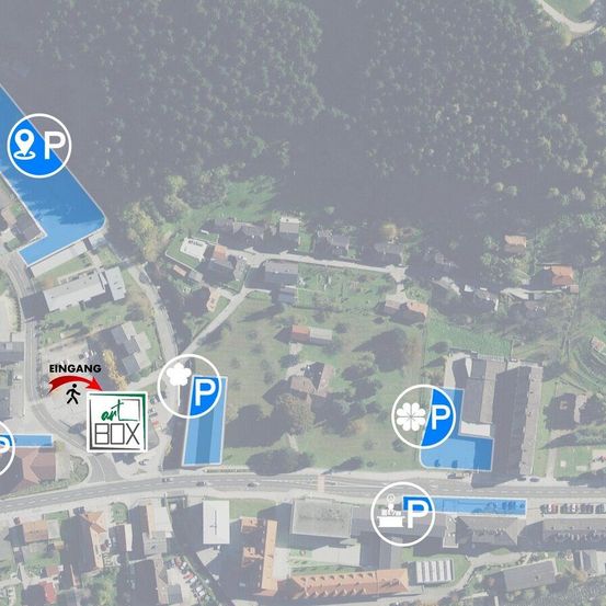 Eine Luftaufnahme zeigt eine kleine Stadt mit mehreren Häusern und Gebäuden. Parkzeichen und eine Box mit der Aufschrift 'art BOX' sind sichtbar. Bäume und Grünflächen umgeben das Gebiet.