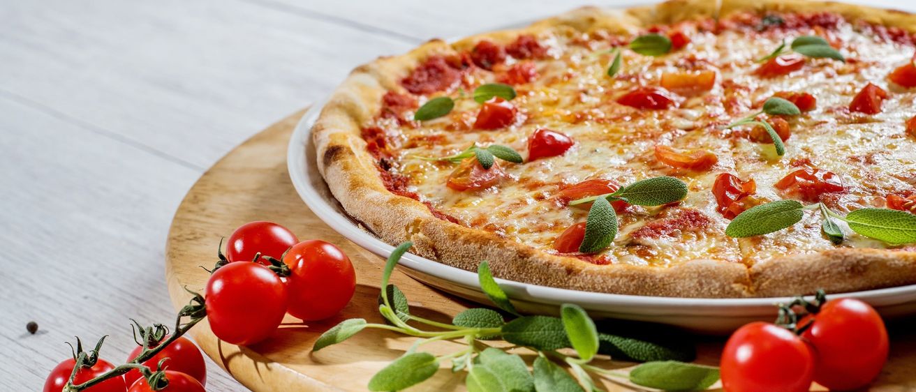Eine ganze Pizza mit Tomatensauce und Käse, garniert mit frischen Tomaten und Salbeiblättern, serviert auf einem Holzteller auf einer weißen Oberfläche.