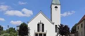 Eine weiße Kirche mit einem grünen Turm und einer Uhr steht in einer sonnigen Landschaft. Die Kirche hat ein Kreuz oben und eine große Tür vorne. Es gibt Bäume und Büsche vor der Kirche.