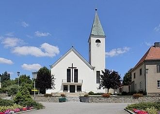 Eine weiße Kirche mit einem Turm und einem Kreuz darauf, umgeben von Bäumen und Blumen, unter einem blauen Himmel mit Wolken.