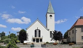 Eine weiße Kirche mit einem Turm und einer Uhr liegt in einer sonnigen Landschaft. Die Kirche hat ein Kreuz auf der Spitze und ein Fenster über dem Eingang. Davor stehen Pflanzen und Bäume.