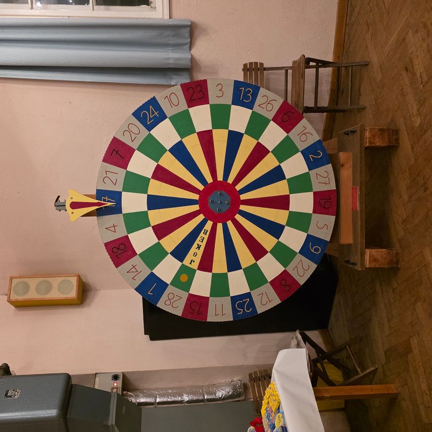 Ein farbenfrohes Drehrad mit Zahlen von 1 bis 36 ist an einer rosa Wand montiert. Drumherum befinden sich Holzstühle, ein hölzerner Schrank und ein Lautsprecher.