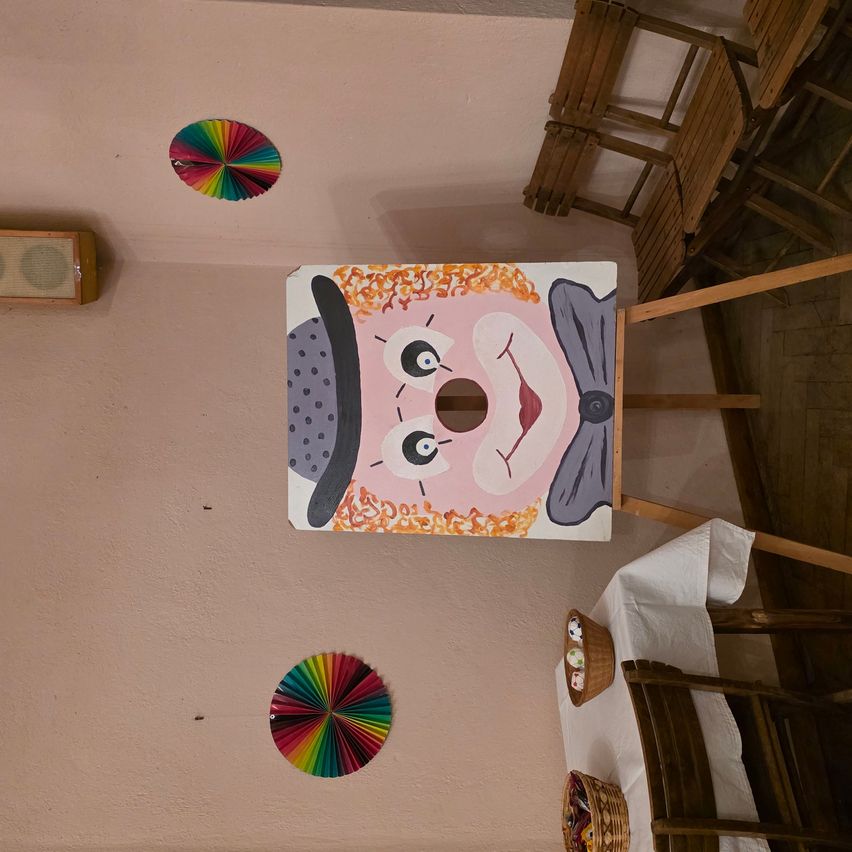 Ein gemalter Clown-Gesicht an der Wand mit einem Loch im Mund. Zwei bunte Windräder und Holzstühle sind ebenfalls vorhanden.